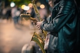 A person wearing a black leather jacket plays a saxophone on a city street at night, with blurred colorful streetlights and people in the background, suggesting a lively urban scene.