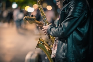 A person wearing a black leather jacket plays a saxophone on a city street at night, with blurred colorful streetlights and people in the background, suggesting a lively urban scene.
