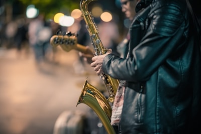 A person wearing a black leather jacket plays a saxophone on a city street at night, with blurred colorful streetlights and people in the background, suggesting a lively urban scene.