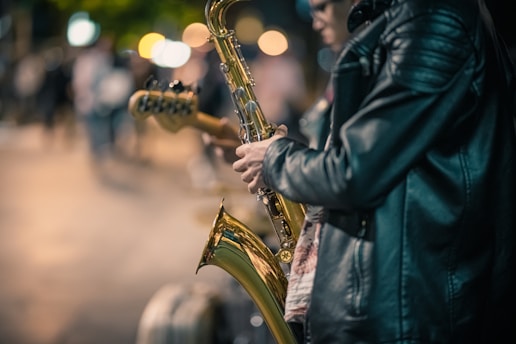 A person wearing a black leather jacket plays a saxophone on a city street at night, with blurred colorful streetlights and people in the background, suggesting a lively urban scene.