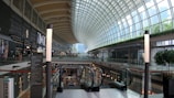 A modern shopping mall with a high, curved glass ceiling allowing natural light to illuminate the space. The mall features multiple levels with various retail stores on each side, and an escalator in the center. There are illuminated signs and vertical columns with lights. The architecture includes sleek, contemporary designs and a blend of greenery.