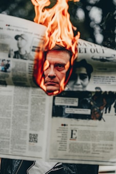 A newspaper held open with a large flame consuming its pages, revealing the face of a man visible through a hole burned in the middle. The background is slightly blurred, emphasizing the fiery foreground.