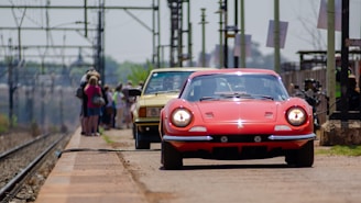 A red vintage sports car and a yellow car are parked on a railway platform with a group of people standing in the background near the train tracks. The platform has an industrial look with overhead power lines and poles.
