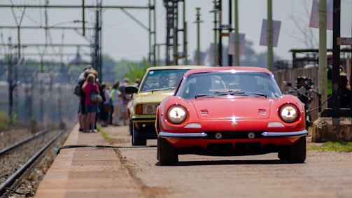 A red vintage sports car and a yellow car are parked on a railway platform with a group of people standing in the background near the train tracks. The platform has an industrial look with overhead power lines and poles.