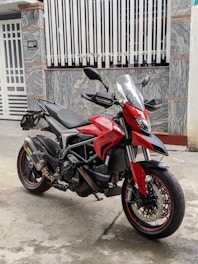 A red and black motorcycle is parked on a concrete floor in front of a textured stone wall and a white gate. The motorcycle has a sleek design with a visible chassis and large disc brakes on the front wheel. It features black handlebars and a transparent windshield.
