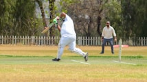 A cricket player dressed in white is captured mid-action, bending forward with a bat poised for a swing on a grassy cricket field. Another player in similar attire stands ready in the background, possibly acting as a wicketkeeper. The field is bordered by a white picket fence with trees providing a leafy backdrop, creating a serene and focused sporting environment.