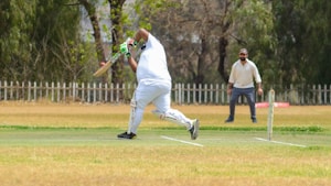 A cricket player dressed in white is captured mid-action, bending forward with a bat poised for a swing on a grassy cricket field. Another player in similar attire stands ready in the background, possibly acting as a wicketkeeper. The field is bordered by a white picket fence with trees providing a leafy backdrop, creating a serene and focused sporting environment.