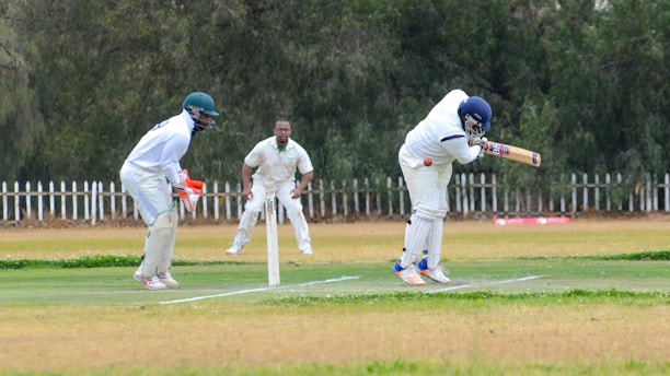 Three cricketers are engaged in a match on a grassy field. Two players are wearing complete protective gear including helmets, pads, and gloves; one is batting while the other is wicketkeeping. A third player is standing upright in a fielding position, wearing a white uniform with a logo. A white picket fence and dense, leafy trees form the background.