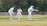 Three cricketers are engaged in a match on a grassy field. Two players are wearing complete protective gear including helmets, pads, and gloves; one is batting while the other is wicketkeeping. A third player is standing upright in a fielding position, wearing a white uniform with a logo. A white picket fence and dense, leafy trees form the background.