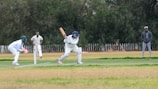 A cricket match scene with a batsman in protective gear preparing to hit a ball. A wicketkeeper crouches behind the stumps, while a bowler and fielders are positioned nearby. The setting is an outdoor field with a grass pitch and trees in the background.