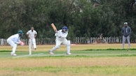 A cricket match scene with a batsman in protective gear preparing to hit a ball. A wicketkeeper crouches behind the stumps, while a bowler and fielders are positioned nearby. The setting is an outdoor field with a grass pitch and trees in the background.
