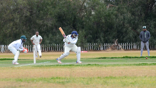 A cricket match scene with a batsman in protective gear preparing to hit a ball. A wicketkeeper crouches behind the stumps, while a bowler and fielders are positioned nearby. The setting is an outdoor field with a grass pitch and trees in the background.