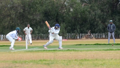 A cricket match scene with a batsman in protective gear preparing to hit a ball. A wicketkeeper crouches behind the stumps, while a bowler and fielders are positioned nearby. The setting is an outdoor field with a grass pitch and trees in the background.