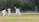 A cricket match scene with a batsman in protective gear preparing to hit a ball. A wicketkeeper crouches behind the stumps, while a bowler and fielders are positioned nearby. The setting is an outdoor field with a grass pitch and trees in the background.