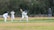 A cricket match scene with a batsman in protective gear preparing to hit a ball. A wicketkeeper crouches behind the stumps, while a bowler and fielders are positioned nearby. The setting is an outdoor field with a grass pitch and trees in the background.