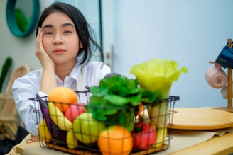 a girl sitting at a table with a basket of fruit and vegetables