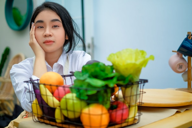 a girl sitting at a table with a basket of fruit and vegetables