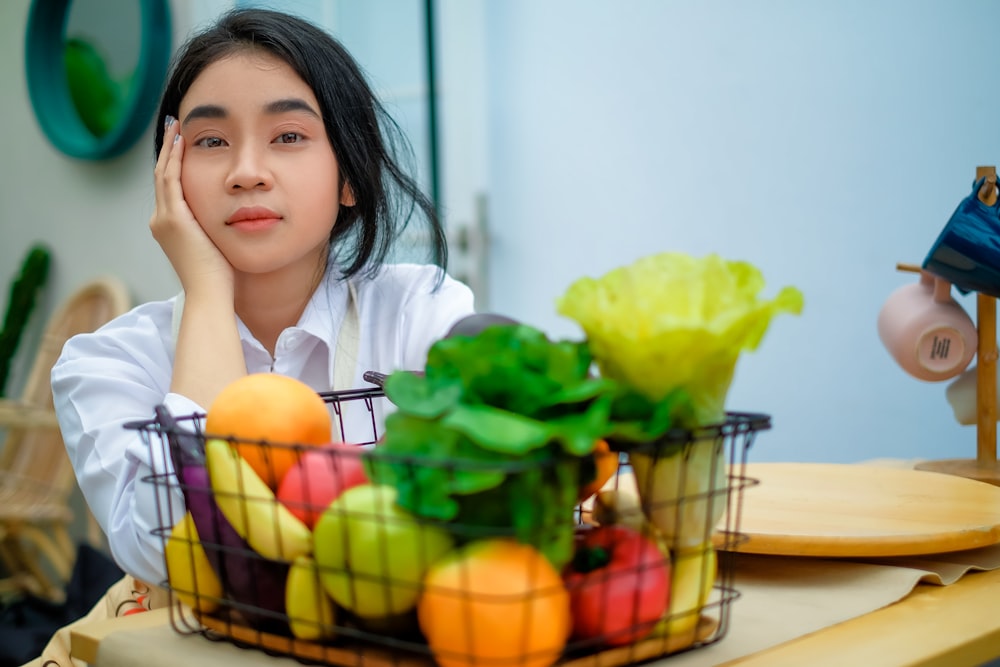 a girl sitting at a table with a basket of fruit and vegetables