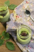 Two small jars filled with green matcha pudding are placed on a wooden table. The pudding is garnished with a sprinkle of green tea powder and what appears to be a berry or red fruit compote. A spoon lies next to the jars on a floral patterned cloth, and several green leaves are placed beside them.
