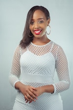 A person is smiling and wearing a white mesh-patterned outfit with hoop earrings. They have dark hair styled to one side and their hands are clasped together.