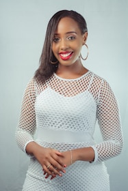 A person is smiling and wearing a white mesh-patterned outfit with hoop earrings. They have dark hair styled to one side and their hands are clasped together.