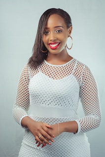 A person is smiling and wearing a white mesh-patterned outfit with hoop earrings. They have dark hair styled to one side and their hands are clasped together.