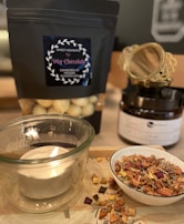 A cozy setup featuring a lit candle in a glass holder, a bowl filled with dried fruit and herbs, and a black package labeled 'My Chocolate' containing white chocolate-covered strawberries. In the background, there's a black jar with decorative gold detailing.