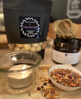 A cozy setup featuring a lit candle in a glass holder, a bowl filled with dried fruit and herbs, and a black package labeled 'My Chocolate' containing white chocolate-covered strawberries. In the background, there's a black jar with decorative gold detailing.