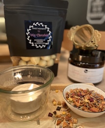 A cozy setup featuring a lit candle in a glass holder, a bowl filled with dried fruit and herbs, and a black package labeled 'My Chocolate' containing white chocolate-covered strawberries. In the background, there's a black jar with decorative gold detailing.