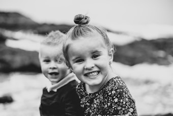 Two young children are captured in a joyful moment, smiling widely. The image is black and white, emphasizing their expressions and the playful atmosphere. The background appears to be an outdoor setting, possibly near rocks or a natural landscape.