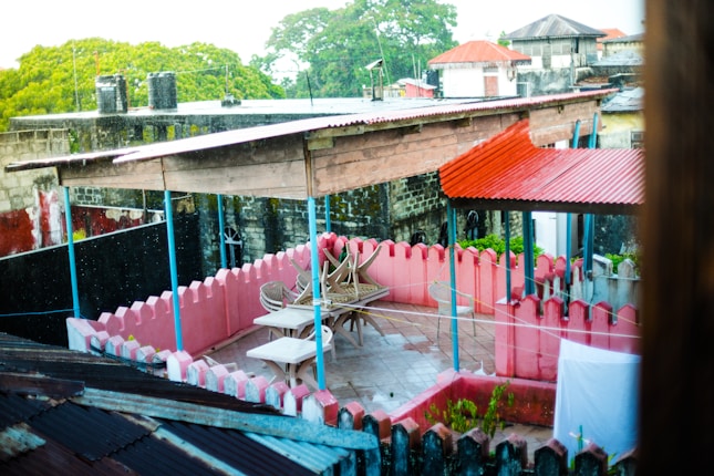 A rooftop terrace with a partially covered seating area featuring tables and chairs. The terrace is enclosed by a low pink wall with a scalloped design. The surroundings include other rooftops with corrugated metal sheets and lush green trees in the background.