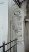 A vertical plaque mounted on a white wall, featuring a list written in the Punjabi language. The plaque is surrounded by architectural details such as carved pillars and is partly covered by a protruding structure. A metallic railing is positioned in front of the plaque.