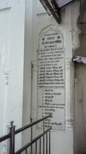 A vertical plaque mounted on a white wall, featuring a list written in the Punjabi language. The plaque is surrounded by architectural details such as carved pillars and is partly covered by a protruding structure. A metallic railing is positioned in front of the plaque.
