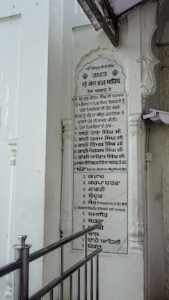 A vertical plaque mounted on a white wall, featuring a list written in the Punjabi language. The plaque is surrounded by architectural details such as carved pillars and is partly covered by a protruding structure. A metallic railing is positioned in front of the plaque.