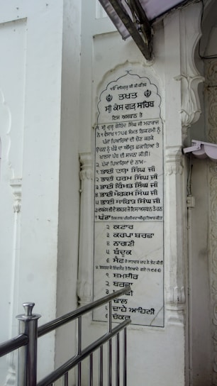 A vertical plaque mounted on a white wall, featuring a list written in the Punjabi language. The plaque is surrounded by architectural details such as carved pillars and is partly covered by a protruding structure. A metallic railing is positioned in front of the plaque.