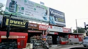 A busy storefront area features multiple signs and advertisements. At the forefront, a large red tractor with the label 'Swaraj 855 FE' is parked. Above the shops, there are prominent billboards for orthopedic capsules and an IVF center. The storefronts include 'Lovely Collection' and 'Shiva Shop' with various products displayed. Coca-Cola branding is visible on signs and awnings. A small motorcycle is parked in front of the shops.
