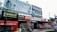 A busy storefront area features multiple signs and advertisements. At the forefront, a large red tractor with the label 'Swaraj 855 FE' is parked. Above the shops, there are prominent billboards for orthopedic capsules and an IVF center. The storefronts include 'Lovely Collection' and 'Shiva Shop' with various products displayed. Coca-Cola branding is visible on signs and awnings. A small motorcycle is parked in front of the shops.