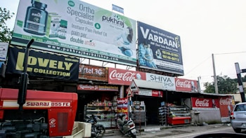 A busy storefront area features multiple signs and advertisements. At the forefront, a large red tractor with the label 'Swaraj 855 FE' is parked. Above the shops, there are prominent billboards for orthopedic capsules and an IVF center. The storefronts include 'Lovely Collection' and 'Shiva Shop' with various products displayed. Coca-Cola branding is visible on signs and awnings. A small motorcycle is parked in front of the shops.