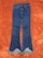 A pair of blue denim jeans with a frayed hem lies flat on an orange textured background. The jeans feature standard pockets and a button closure.