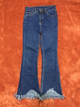 A pair of blue denim jeans with a frayed hem lies flat on an orange textured background. The jeans feature standard pockets and a button closure.