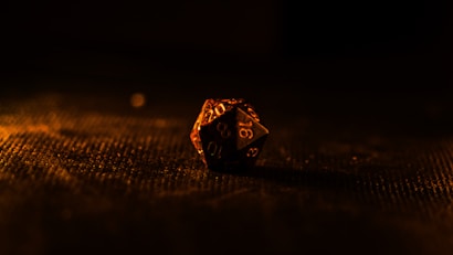 A single twenty-sided die sits on a textured surface, illuminated by warm, low lighting that casts dramatic shadows. The background is mostly dark, emphasizing the die's intricate numbers and facets.