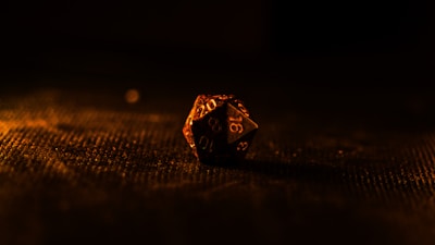 A single twenty-sided die sits on a textured surface, illuminated by warm, low lighting that casts dramatic shadows. The background is mostly dark, emphasizing the die's intricate numbers and facets.