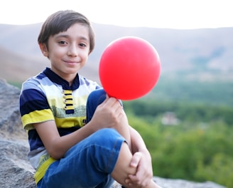 a young boy sitting on a rock holding a red balloon