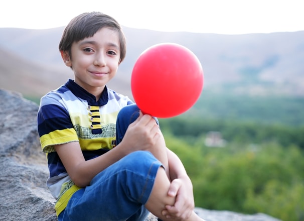 a young boy sitting on a rock holding a red balloon