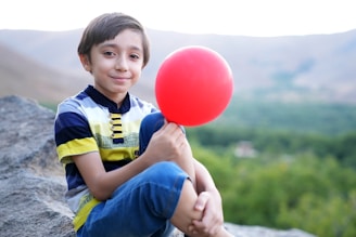 a young boy sitting on a rock holding a red balloon