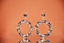 A pair of large circular hoop earrings adorned with various gemstones. The gemstones are a mix of blue and clear hues, set in a gold framework. The earrings are placed on a reflective surface with an orange background, creating a warm and elegant atmosphere.