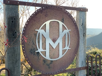 A large, round, rusted metal sign with a modern, silver monogram in the center, mounted on wooden posts. The background features barren branches and a distant view of hills under clear skies.