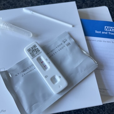 A COVID-19 rapid lateral flow test kit is displayed, including a test cassette with a visible test line and control line, a sealed swab, and NHS Test and Trace paperwork.