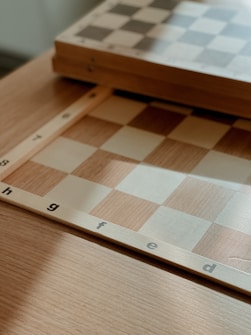 A close-up view of a wooden chessboard with light and dark squares. The board is positioned on a wooden table, and visible letters along the edge indicate columns. The lighting casts soft shadows, giving a warm and cozy atmosphere to the scene.