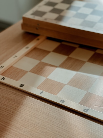 A close-up view of a wooden chessboard with light and dark squares. The board is positioned on a wooden table, and visible letters along the edge indicate columns. The lighting casts soft shadows, giving a warm and cozy atmosphere to the scene.
